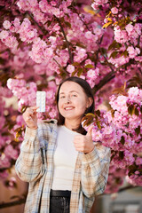 Fototapeta premium Woman allergic suffering from seasonal allergy at spring. Young happy woman showing allergy drugs, giving thumbs up, posing in blossoming garden at springtime. Antihistamine medication concept