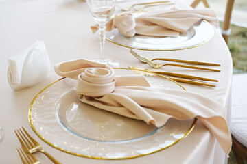 Elegant table setting with gold cutlery, glassware, and folded napkins on white tablecloth, featuring clear plates with gold rims and decorative elements arranged neatly