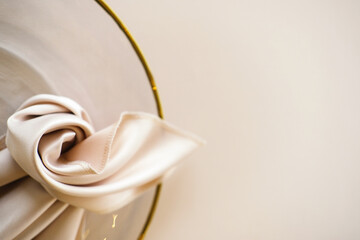 Wedding banquet table setting with white satin napkin tied in a knot on a gold-rimmed plate against a light beige background with copy space