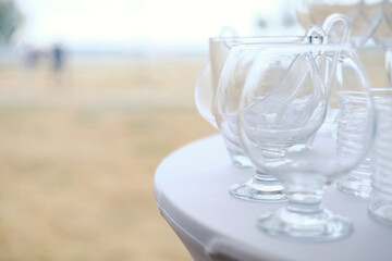 Clear glass cups arranged on white tablecloth with blurred background of outdoor setting and people walking in distance during daytime