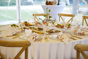 Round dining table set with white tablecloth, plates, cutlery, glasses, napkins, and various food items arranged in a tent with greenery visible outside