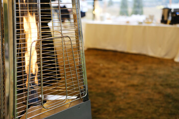 Outdoor patio heater with flame visible in foreground, blurred table with white tablecloth and dishes in background, grass surface beneath, evening setting with soft lighting