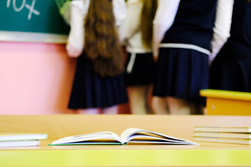 Open book on wooden desk in classroom with blurred students in black skirts and white blouses standing near green chalkboard during lesson time
