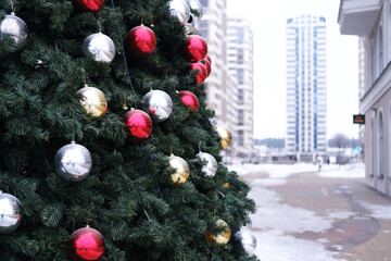 Christmas tree with green branches and red, silver, and gold ornaments in foreground with buildings and snow-covered ground in background during winter season