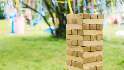 Wooden block tower stacked vertically on green grass with colorful ribbons hanging from tree branches in background during outdoor event with blurred objects nearby