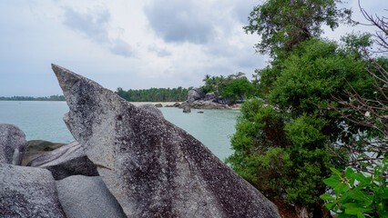 Distinctive sharp granite rock formation on a tropical rocky coastline with calm sea, green vegetation, and cloudy sky.