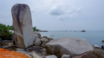 Tall standing granite boulder on a tropical rocky beach with red sand foreground, calm sea, and cloudy sky.