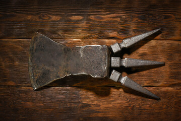 Forged medieval axe head with three spike tangs placed horizontally on dark wooden plank workbench table background.