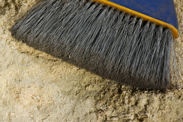Hand broom with dense gray bristles cleaning fine sawdust on workshop surface background.