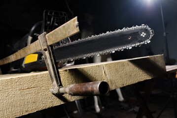 Chainsaw fixed with metal clamp on wooden beam during woodworking process in dark workshop environment background close up.