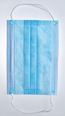 A close-up vertical studio shot of a disposable light blue pleated surgical face mask against a white background.