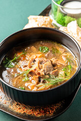 A hearty bowl of mushroom soup with dill and parsley is served with pieces of fresh bread and a side of sour cream on a black plate set on a dark green tablecloth