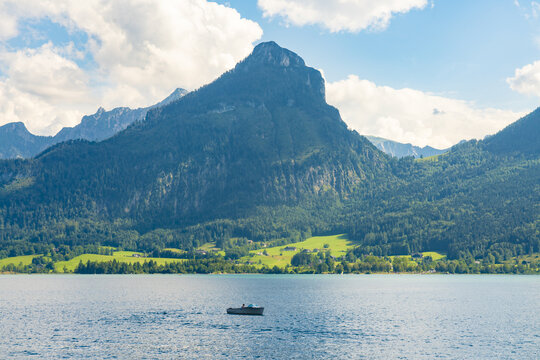 View of a solitary boat glides on the lake's serene surface, reflecting the towering, forest-clad mountains under a vast sky, Wolfgangsee, Salzkammergut, Austria.