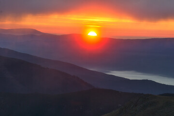 View of a fiery sun melting into the horizon over the misty, layered mountain ridges, casting a warm glow across the serene landscape, Skalka, Banska Bystrica Region, Slovakia.