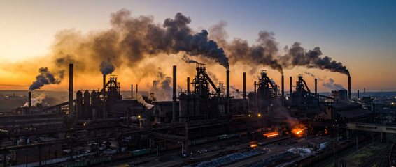 Fototapeta premium Aerial view of a sprawling industrial steel mill with smoking chimneys silhouetted against a dramatic sunset sky. Heavy industry, energy production, and air pollution