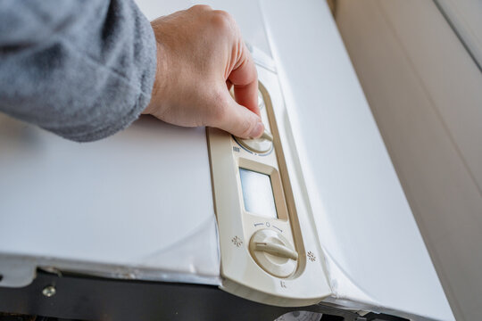 Hand regulating the temperature dial of a wall-mounted boiler with visible pipes underneath, illustrating residential heating control, energy efficiency and daily home maintenance tasks.