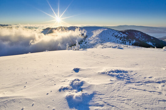 View of a sunburst over snow-covered peaks and fluffy clouds, with snow drifts in the foreground creating a serene winter landscape, Kosarisko, Banska Bystrica region, Slovakia.