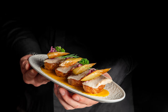 waiter holding a grey oval plate with several pieces of elegant fish canap&eacute;s or appetizers garnished with pear slices, red onion, and parsley on a yellow sauce, against a black background
