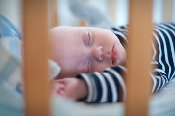 An infant is peacefully asleep in a crib. The baby wears a striped shirt and is resting on a white sheet, behind the wooden bars of the crib. The afternoon light is soft.