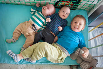 Three young brothers are laying in their baby bed. Two are infants and one is a young boy. They are wearing casual clothing and socks. One boy smiles while holding a stuffed animal.