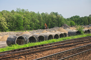 Concrete pipes and gravel piles line a construction site beside active railway tracks, excavator working near forested backdrop. Scene blends transportation infrastructure with utility development