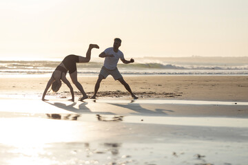 Couple practicing capoeira movements on sandy beach at sunset