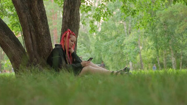 individual relaxes with journal under tree, serene scene of woman immersed in creative writing session, peaceful park setting showing woman with red dreadlocks engaging in quiet composition