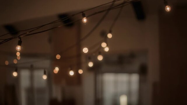 Shifting focus, camera moving across string of warm filament bulbs in large room, revealing sockets