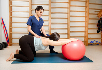 Pregnant woman doing prenatal exercises with physiotherapist using a fitness ball. Female specialist guiding pregnant client in maternity physical therapy session.