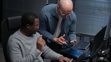 Two multiethnic colleagues collaborating in dark office using digital tablet to analyze data and perform cybersecurity tasks on network and systems - Powered by Adobe