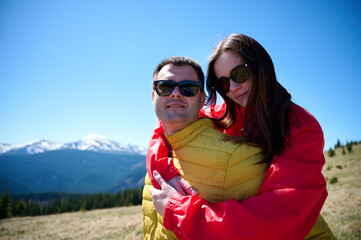 Happy couple enjoying mountain adventure. Man carrying smiling woman on his back. Outdoor romance, hiking and travel in the mountains on a sunny day. Love and fun in nature.