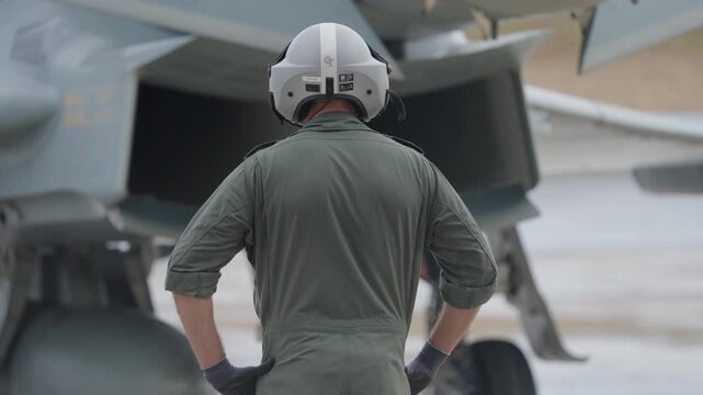 Rear view of a ground crew member in helmet and uniform standing near a fighter jet engine intake during preflight inspection on airbase apron
