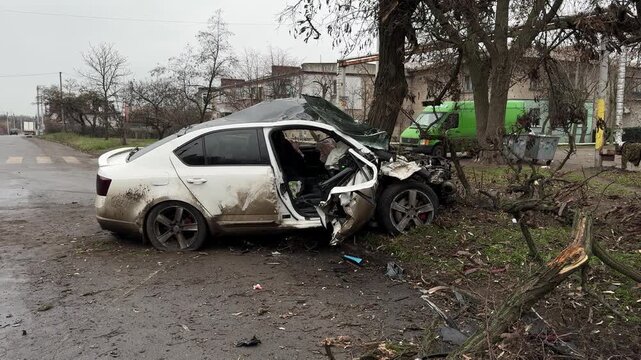 A passenger car sustains extensive damage after a serious traffic accident. The car crashed into a tree on the side of the road, completely destroying the front end, shattering the windshield,