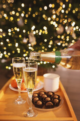Pouring champagne into glasses against backdrop New Year tree with Christmas lights. Celebrating Christmas and New Year's with friends in evening, corporate event. Romantic, elegant atmosphere couple