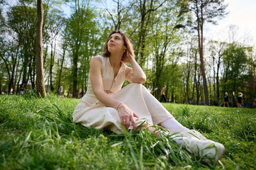 Relaxed woman sitting on green grass in a spring park, enjoying warm sunlight and nature. Peaceful lifestyle moment, outdoor leisure, mindfulness, and natural happiness concept.