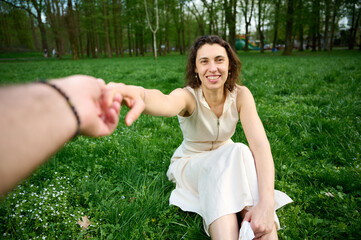 Smiling woman sitting on green grass in a park, reaching out and holding hands with partner. Romantic spring moment, connection, trust, love, outdoor lifestyle and happiness concept.