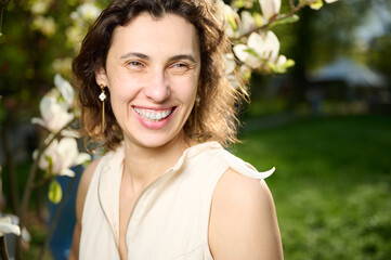 Smiling woman portrait outdoors near blooming magnolia flowers in spring garden. Natural beauty, fresh skin, and joyful expression in soft sunlight. Concept of spring and harmony with nature