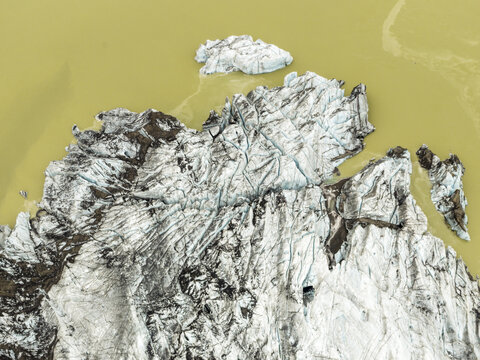 Aerial view of glacial ice, carved by time and weather, sits starkly against the pale yellow waters of Solheimajokull, Myrdalshreppur, Iceland.