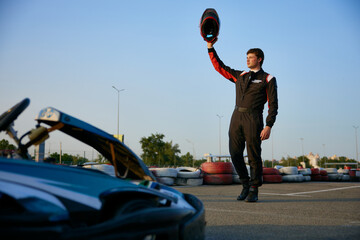 A kart racer stands proudly on the track, raising his helmet in a victorious gesture