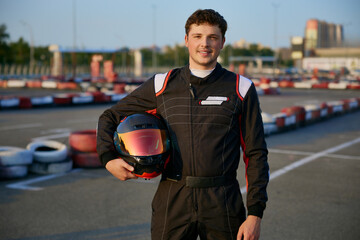 A confident go-kart racer stands in full gear, holding a helmet in a vibrant outdoor circuit