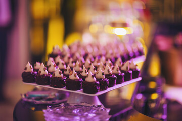Small brownie bites are neatly placed on a white serving tray at a festive event. Each bite has cream topping and is surrounded by soft lighting that adds warmth to the space