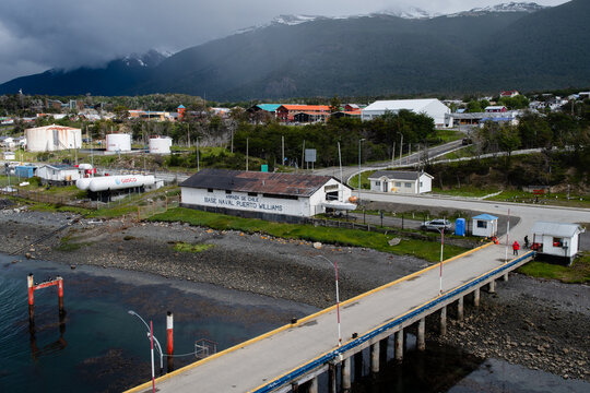 Aerial view of a naval base and pier stretching into the dark water, contrasting with the bright buildings and the snow-capped mountains under a cloudy sky, Puerto Williams, Chile.