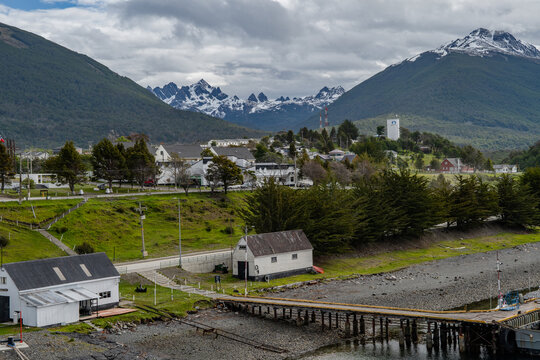 Aerial view of a quaint town nestled between verdant hills and snow-capped Dientes de Navarino mountains, where a wooden pier stretches into the water, Puerto Williams, Chile.