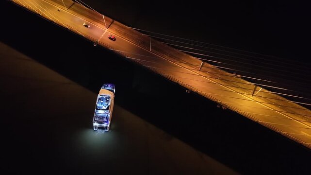 Top down night drone shot showing an illuminated boat passing beneath San Roque Gonz&aacute;lez de Santa Cruz Bridge, with moving cars visible on the bridge above