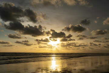 Golden sunset over a tropical beach with dramatic clouds and sun rays reflecting on the wet sand and waves.