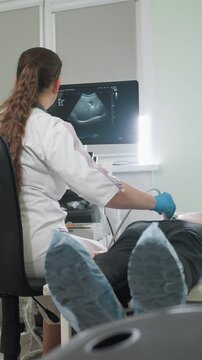 female ultrasound technician scanning patient abdomen bedside for liver and gallbladder assessment, gloved hand moves transducer while monitor displays live images, patient lies on exam bed with blue