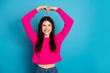Young woman in pink sweater forms a heart with her arms against a blue background