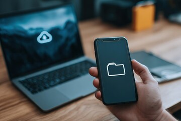 Smartphone displaying folder icon in hand, with laptop featuring cloud symbol on wooden desk, illustrating digital storage and technology concepts in modern workspace