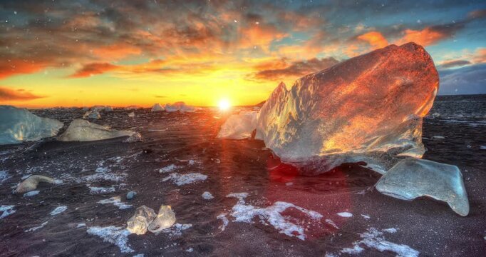 Static slow motion shot of sunset at Diamond Beach in Iceland with gently falling snow. Glowing ice crystals on black sand create a calm, cinematic winter atmosphere.