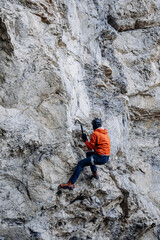 Male Dry Tooling Climber In Red Jacket On Steep Rock Face: Technical Ascent Using Ice Axes, Crampons, And Rope On Vertical Limestone Cliff